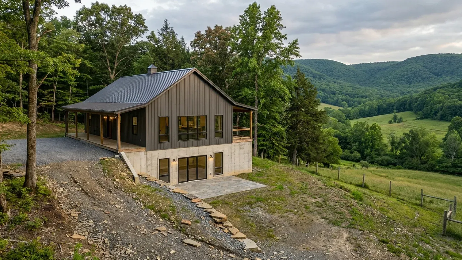 A barndominium with a walkout basement built into a sloping lot, the lower level opening onto a patio at grade, with forested ridges behind.